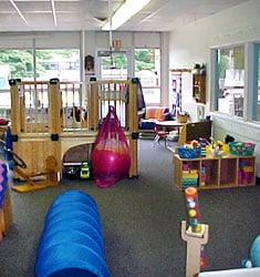 A child care room at the center filled with toys, a wooden play structure and lots of natural light coming from a wall of windows at the back of the room