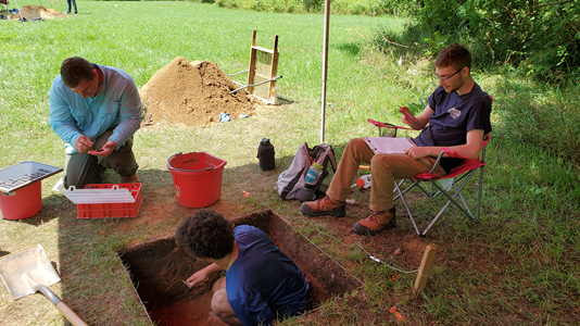 Students working at an archaeological dig under a tent. One crouched in a square hole with carious buckets and tools surrounding.