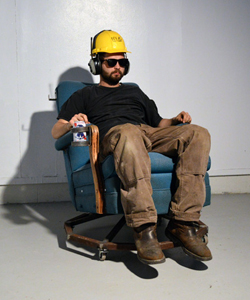 Portrait of Dane Winkler in a hard hat and ear muffs, sitting on a modified arm chair with a drink in one hand