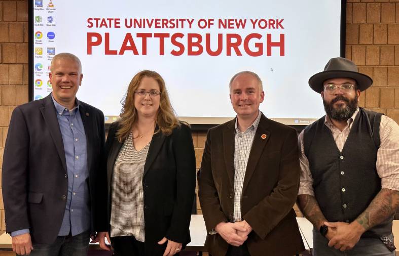 Four adults standing shoulder-to-shoulder in front of a projection of the SUNY Plattsburgh logo.