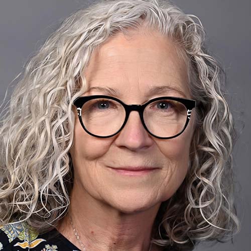Woman with shoulder length, curly white hair wearing black glasses smiles without teeth.