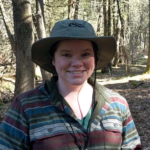 Woman standing outside in the fall, surrounded by trees, wearing a bucket hat and flannel shirt.
