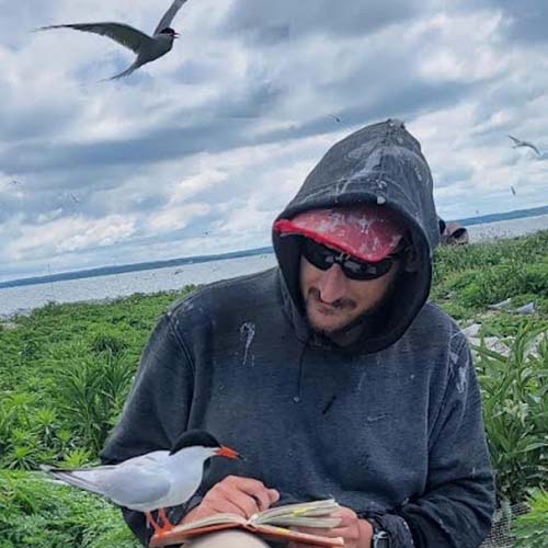 Man sitting by a body of water with a dark hooded sweatshirt and baseball cap; a sea bird is curiously perched on his notebook.