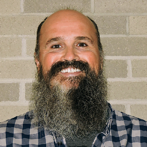 A man wearing a flannel shirt with a bushy black and gray beard stands, smiling brightly, against a light brick wall