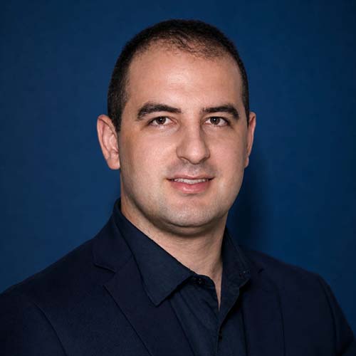 Man with dark hair wearing a black shirt stands against a dark blue background for a headshot.