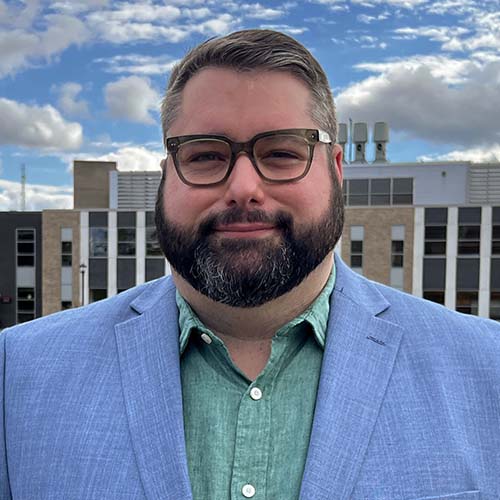 Man with dark brown glasses and blue sports coat stands outside in front of an academic building. Blue sky peppered with white clouds in the background.