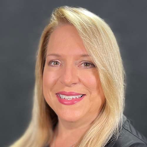 Woman with long blond hair in a dark dress shot against a dark background. She smiles for the camera