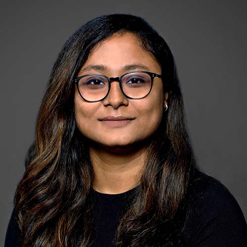 Woman with long dark hair and a dark shirt against a dark background sits for a headshot.