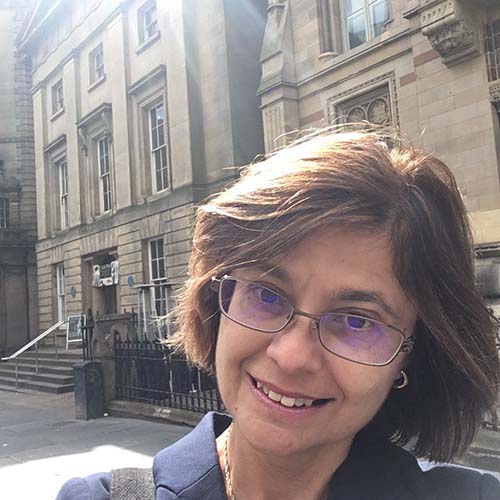 Woman on a city street, sun shining behind her, takes a selfie in front of historic residential buildings