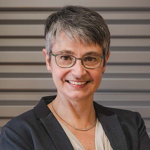 Woman with a salt-and-pepper colored pixie-cut wearing glasses and professional attire smiles for the camera