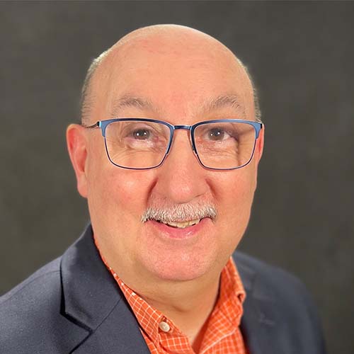 Man with blue-rimmed glasses, orange collared shirt and blue shirt jacket smiles brightly for the camera.