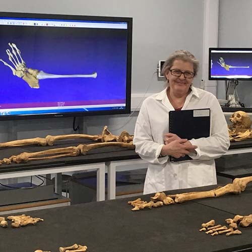 Woman with glasses, a lab jacket and clipboard, and short gray hair stands in front of a table with a skeleton sprawled out.