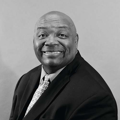 Black and white image of a man wearing a dark suit and neck tie, sitting for a professional photo