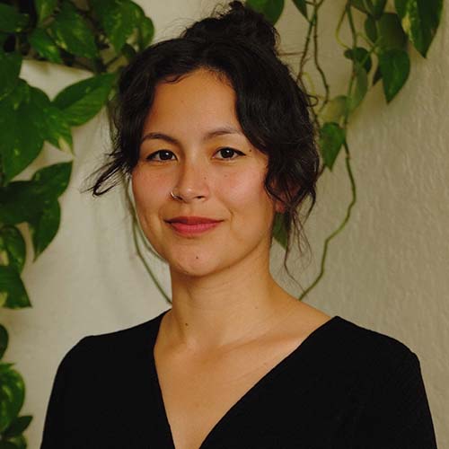 Close-up of a woman with wavy dark hair pulled into a messy bun stands against a textured wall, a pothos climbing the wall behind her.