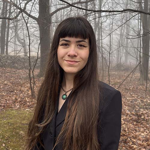 A person with long, dark hair stands outside on a foggy late fall day, with bare trees in the background.