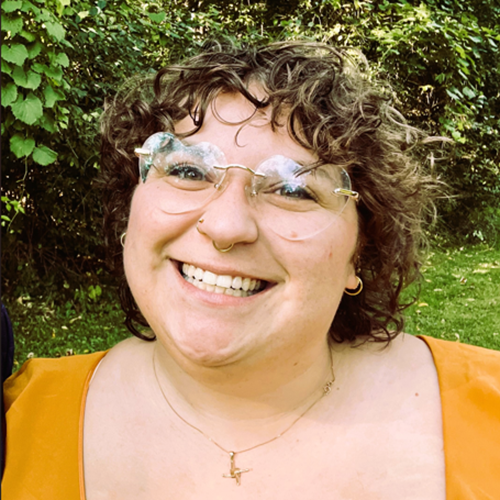 Person with short curly hair stands outside against greenery, smiling brightly for the camera. They wear heart-shaped eye glasses.