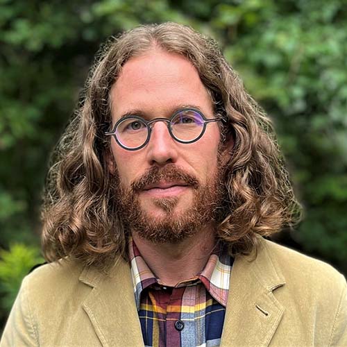 Man with shoulder-length curly hair parted down the middle stands outside. He's got round blue eyeglasses and wears a camel-colored corduroy jacket.