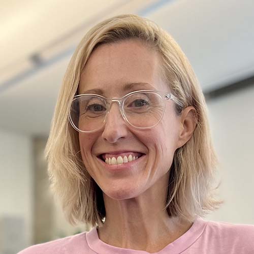 Woman with clear frame glasses and blond shoulder-length hair smiles widely for the camera