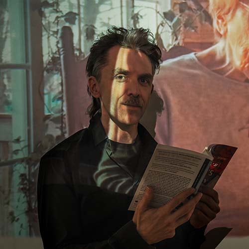 man standing against a projection of an old woman reading, while holding a book himself