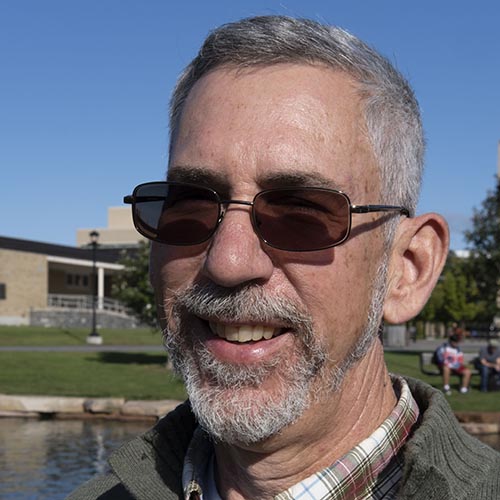 Man with plaid shirt and earthy-green sweater smiles for the camera on a sunny, blue-skied day.