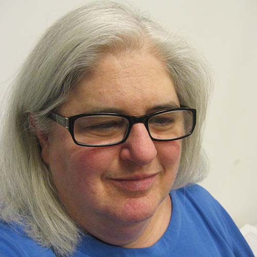 Woman with long white hair and black rectangular glasses shot close up, head tilted down, shot from above.