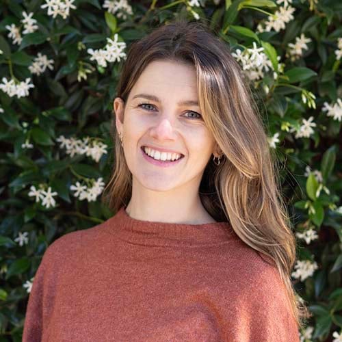 Woman stands outside against a flowering hedge, smiling for the camera