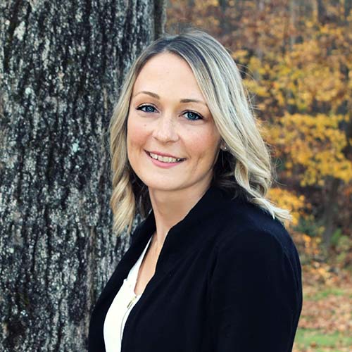 Woman in professional clothing stands outside against a tree, fall foliage around, posed for a headshot