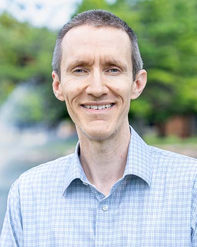 Man wearing a plaid button-up shirt stands outside for a professional headshot