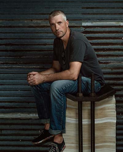 man in an industrial-looking studio sitting on a stack of what appears to be newspapers