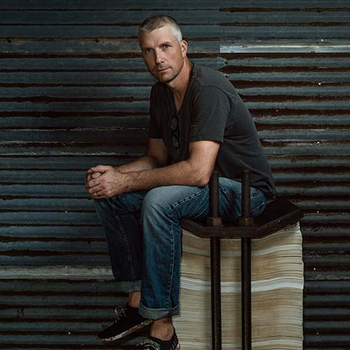 Man, against an industrial corrugated metal background sits atop a high stack of papers