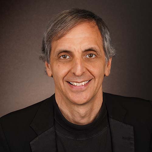 professional headshot of a man wearing a black sweater and tuxedo jacket, smiling widely for the camera