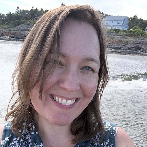 Close-up of a woman with light blue eyes and wavy brown hair wearing a sleeveless flowery shirt at what appears to be coastal Maine beach.