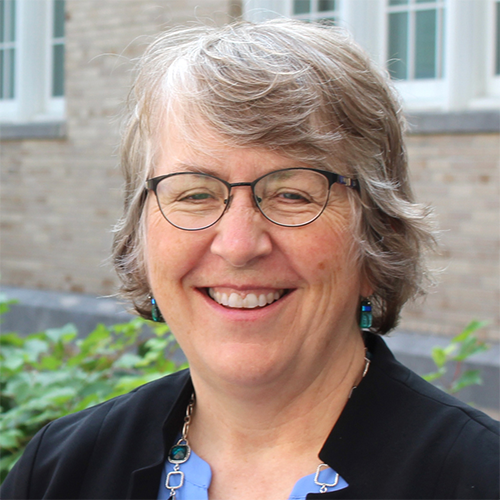 Woman with short gray hair stands outside in front of a stone building, smiling for a headshot