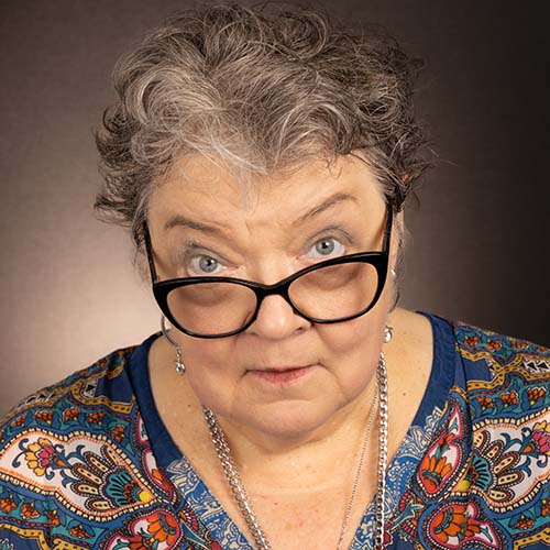 Professional headshot of woman with short, curly, salt-and-pepper hair looking up slightly at the camera with glasses down on the bridge of her nose.