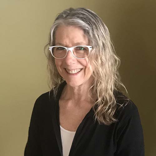 Woman with long blond waivy hair stands against a mustard-gold wall smling brightly. She wears white cat eye glasses.