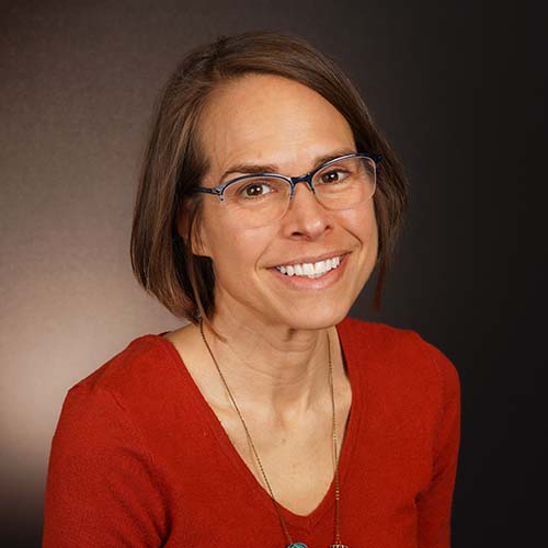 Woman with chin-length hair sits for a professional headshot. She wears an earthy red v-neck knit shirt.