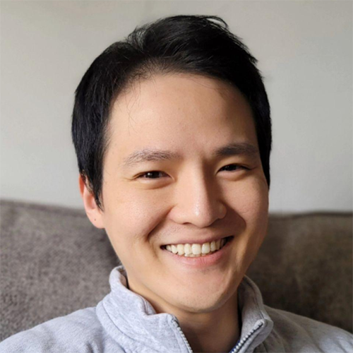 Man with a sweater on sits easily on a couch, smiling brightly.