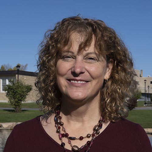 Woman with curly brown hair and dark chunky necklace poses outside during a blue-sky bright day.