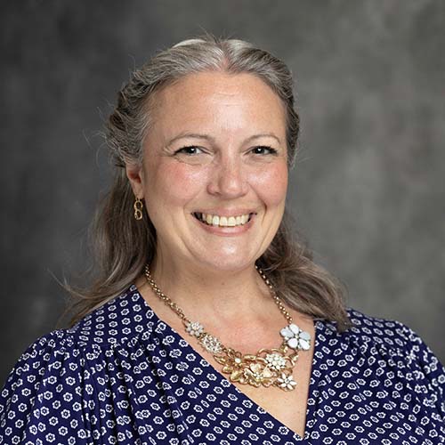Person with long brown and gray hair pulled away from their face, smiling brightly with a chunky gold necklace.