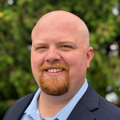 Man with blue suit jacket stands outside against greenery for a headshot.