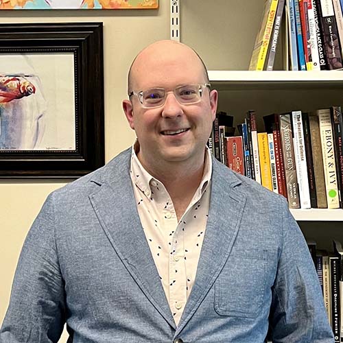 Man with a blue cardigan stands in an office in front of a bookshelf