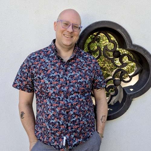 Smiling man, hands in pocket, with a multi-color short-sleeve shirt, stands outside in front of a clover-shaped window