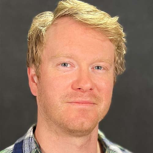 Man with blondish-red hair and blue plaid shirt smiles for the camera with closed lips