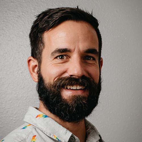 A man with dark hair and dark beard sits at a slight angle, smiling for the camera. He is wearing a light button up shirt with rainbow lightning bolts on it.