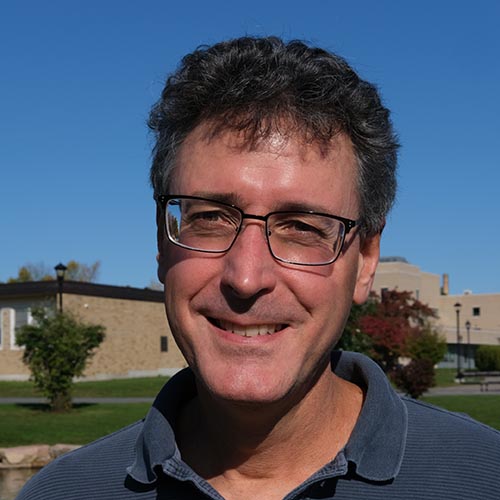 Man with blue polo shirt smiles for the camera while outside on a sunny, blue-skied day.