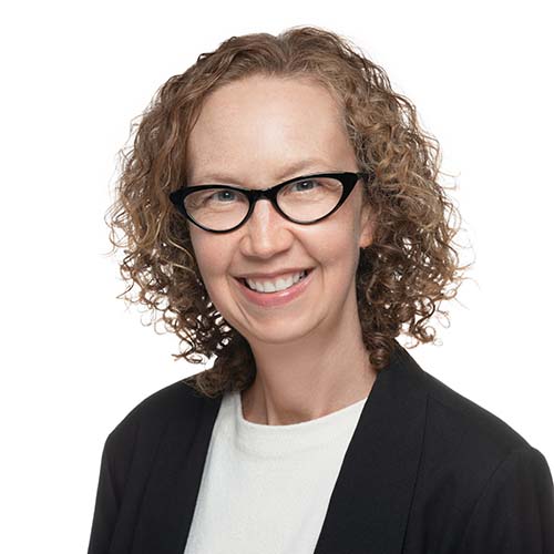 Slight side profile of a woman with light brown curly hair and cat-eye glasses. She's wearing a black blazer and white shirt.