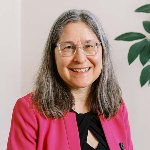 Woman with long, gray hair sits for a portrait. She wears a black top and fuchsia jacket.