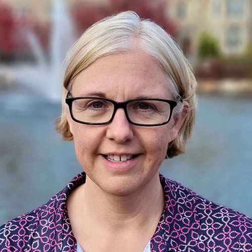 Woman with a graying-blonde bob stands in front of the Hawkins Hall pond fountain. She smiles for the camera brightly.