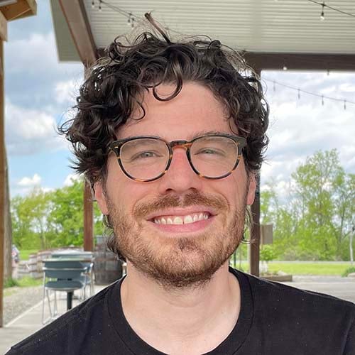 Man with curly dark hair wearing round tortoise shell glasses sits outside, smiling for picture.