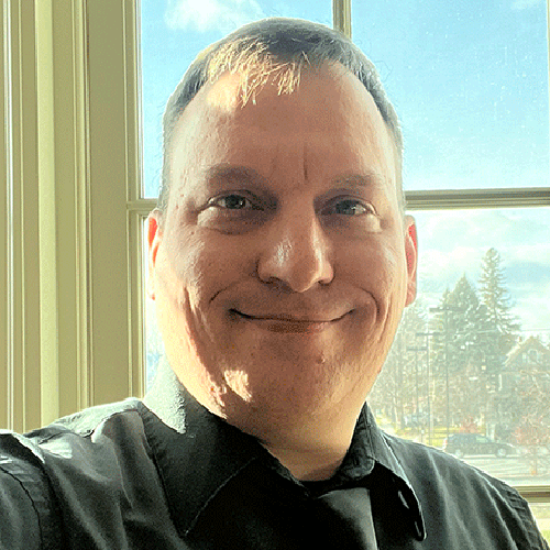 Matthew Pray, wearing a black dress shirt and black tie, backlit by a sunny window.